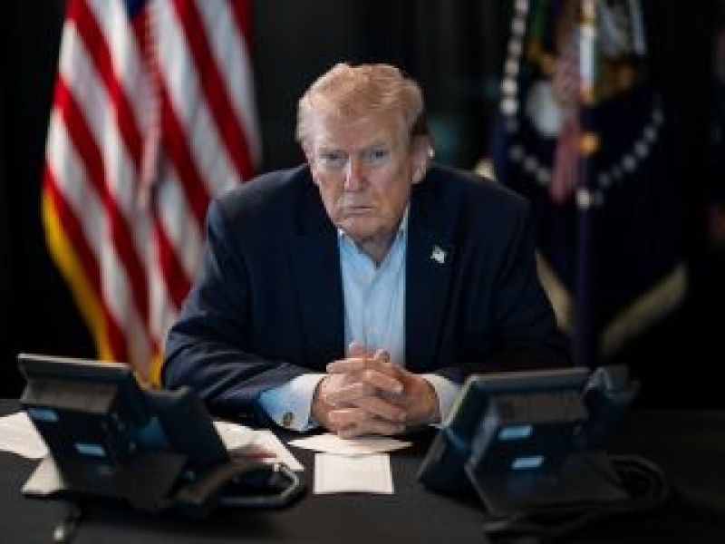 Serious-looking Donald Trump sits at desk, hands folded,in front of comms equipment with American flag in background