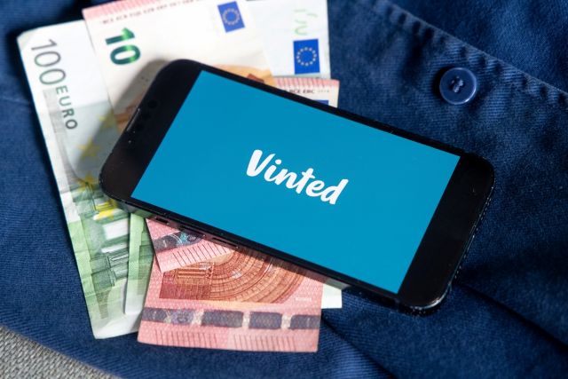 Un téléphone portable affichant le logo Vinted à l'écran, sous une pile d'euros et une chemise en jean bleu.