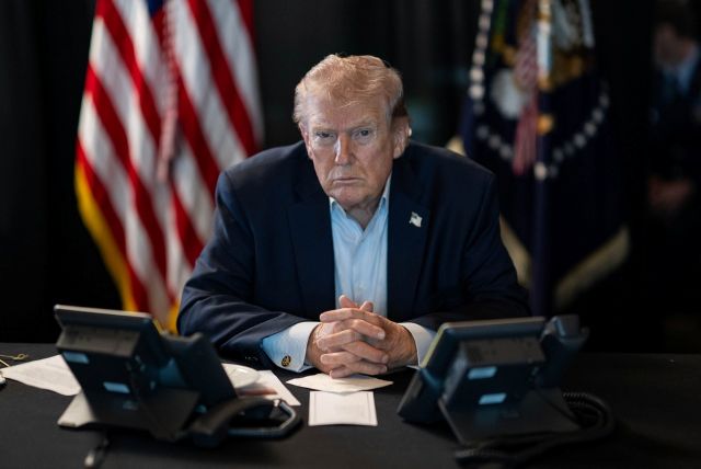 Serious-looking Donald Trump sits at desk, hands folded,in front of comms equipment with American flag in background