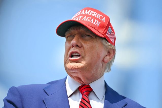 Donald Trump portant une casquette rouge de MAGA, un costume bleu et une cravate rouge, sur fond de ciel bleu.