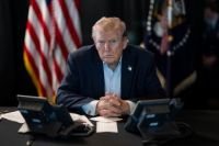 Serious-looking Donald Trump sits at desk, hands folded,in front of comms equipment with American flag in background