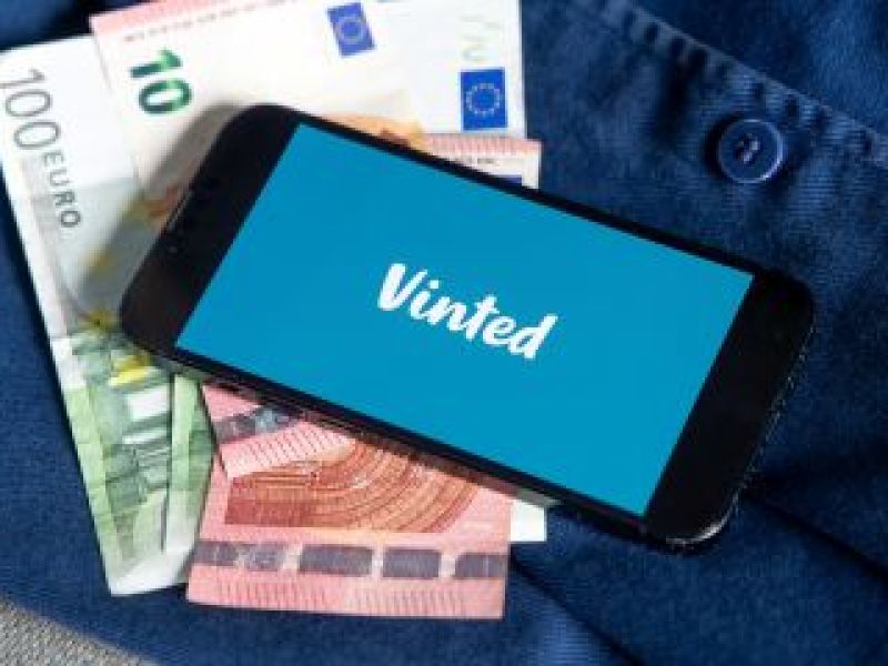 Un téléphone portable affichant le logo Vinted à l'écran, sous une pile d'euros et une chemise en jean bleu.