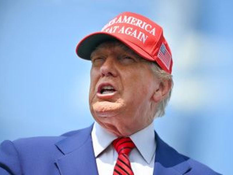 Donald Trump portant une casquette rouge de MAGA, un costume bleu et une cravate rouge, sur fond de ciel bleu.