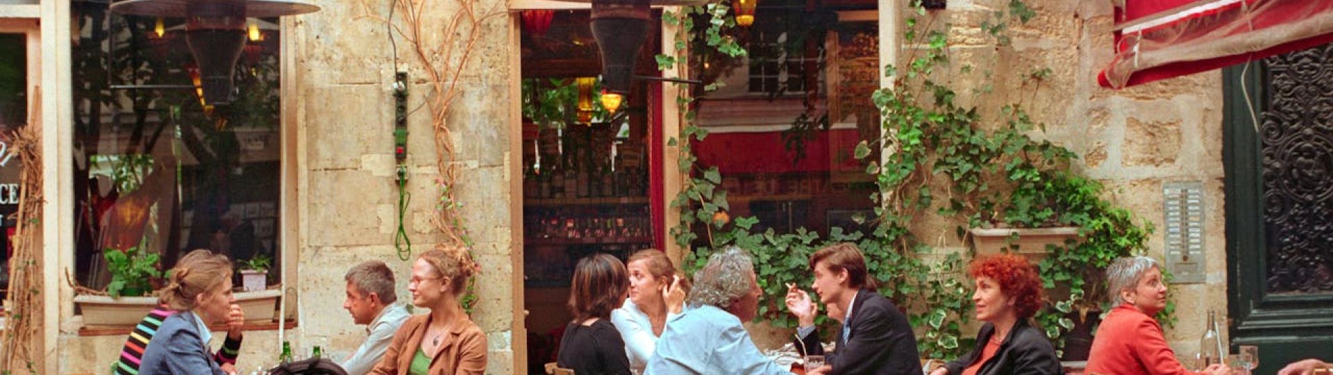 A group of people sat outside of a French cafe business with plants and vines in the background